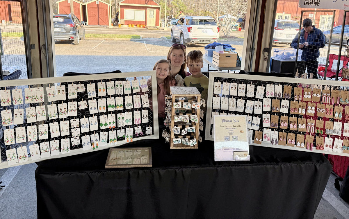 Erin and her two children behind the Bloomin' Bees Clay Co. booth at a Virginia Beach market, with two display boards of handmade polymer clay earrings
