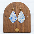White and blue floral teardrop polymer clay dangle earrings displayed on a wooden earring stand